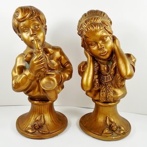 May include: Two gold-colored busts of children. One plays a saxophone, the other covers their ears. Both are on pedestals with floral accents. The statues are likely made of a resin or plaster material.