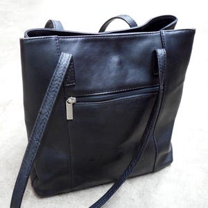 May include: A black leather tote bag with two short handles and two long shoulder straps. The bag has a zippered pocket on the back and a silver zipper pull. The bag is rectangular and appears to be empty.