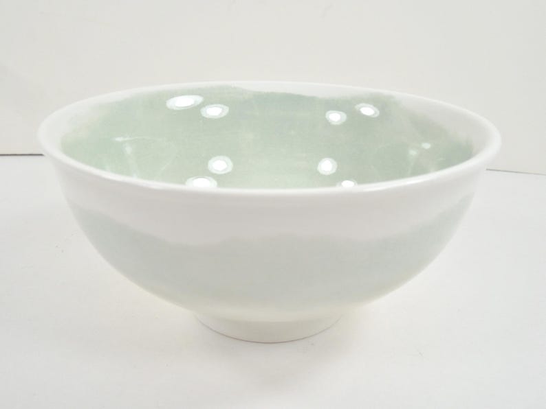 May include: A white ceramic bowl with a pale green interior. The bowl has a rounded shape and a smooth, glossy finish. The interior has a subtle, speckled pattern. The bowl is suitable for serving food.