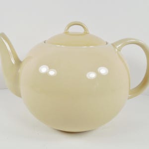 May include: A cream-colored ceramic teapot with a round body and a lid. The teapot has a spout and a handle.