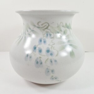 May include: A white ceramic vase with a rounded body and a flared rim. The exterior features a hand-painted design of delicate, light blue floral clusters and soft green foliage vines that wrap around the upper portion of the vessel.