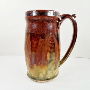 May include: A tall ceramic mug featuring a glossy glaze with a deep brown rim that transitions into a warm amber and tan base. The surface displays vertical, textured brushstroke patterns throughout the body and a curved, sturdy handle on the side.