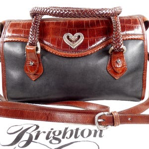 May include: A two-tone handbag featuring a black leather body and brown crocodile-embossed leather trim. It includes braided leather handles, a detachable shoulder strap with a silver-tone buckle, and a central silver-tone heart-shaped emblem.