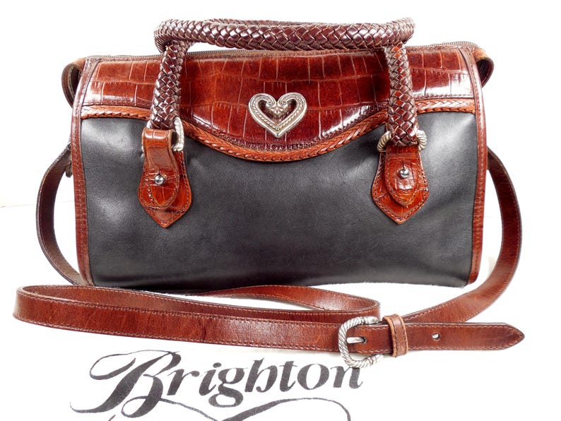 May include: A two-tone handbag featuring a black leather body and brown crocodile-embossed leather trim. It includes braided leather handles, a detachable shoulder strap with a silver-tone buckle, and a central silver-tone heart-shaped emblem.