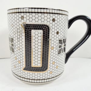 May include: White ceramic mug with a black handle and the letter "D" in a mosaic design. The mug features a honeycomb pattern with black and gold accents. A decorative floral design is also present.