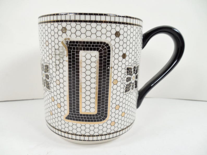 May include: White ceramic mug with a black handle and the letter "D" in a mosaic design. The mug features a honeycomb pattern with black and gold accents. A decorative floral design is also present.