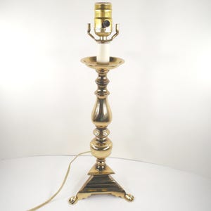 May include: A vintage brass table lamp with a candlestick design. The lamp features a gold-colored finish, a bulb socket, and a white candle-shaped light bulb. The base is square with decorative feet. The lamp is approximately 18 inches tall.