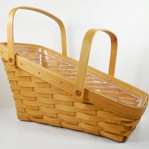 May include: A light brown, woven picnic basket with a clear plastic insert and two arched handles. The basket has a slanted design and is made of woven wood strips.
