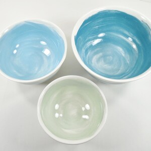 May include: Three ceramic bowls with white exteriors and colourful interiors. The bowls are in shades of blue and green, with a swirled paint effect. The bowls vary in size, with the largest bowl at the top and the smallest at the bottom.