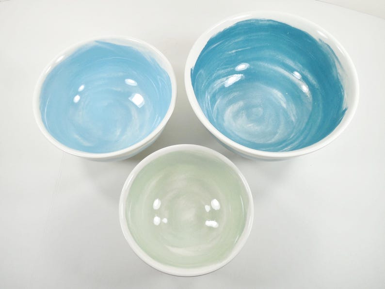 May include: Three ceramic bowls with white exteriors and colourful interiors. The bowls are in shades of blue and green, with a swirled paint effect. The bowls vary in size, with the largest bowl at the top and the smallest at the bottom.
