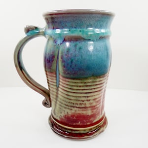 May include: A tall ceramic mug featuring a flared rim and a curved handle. The upper portion has a glossy blue and purple glaze, while the lower section displays horizontal ridges with a blend of red, tan, and brown earthy tones throughout.