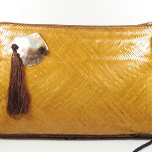 May include: A rectangular, woven straw handbag with a zipper closure and a dark brown rope strap. The bag features a decorative seashell and a brown tassel. The bag is a light golden color.