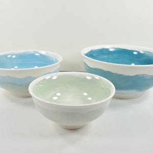 May include: Three ceramic bowls of different sizes. The bowls have a white exterior and a painted interior. The largest bowl has a blue interior, the medium bowl has a green interior, and the smallest bowl has a light blue interior.