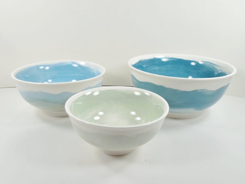 May include: Three ceramic bowls of different sizes. The bowls have a white exterior and a painted interior. The largest bowl has a blue interior, the medium bowl has a green interior, and the smallest bowl has a light blue interior.
