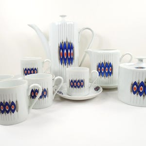 May include: A white ceramic coffee set with a blue, red, and gold geometric design. The set includes a coffee pot, creamer, sugar bowl, cups, and saucers. Vertical lines and a repeating pattern decorate the pieces.
