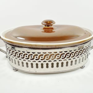 May include: Oval brown ceramic casserole dish with a lid and a silver-colored metal stand. The dish has a white rim and a brown knob on the lid. The stand has decorative cut-out patterns and curved handles.