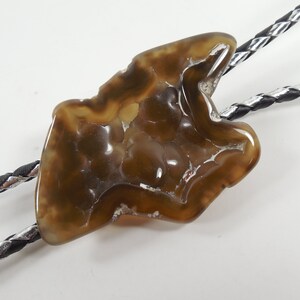 May include: A brown agate bolo tie with a silver and black braided cord. The agate is a natural stone with a unique pattern.