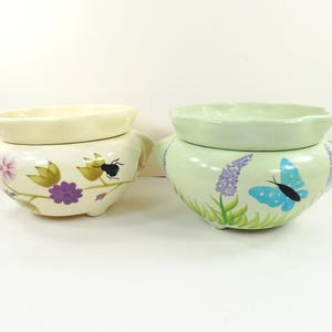 May include: Two ceramic planters with scalloped rims and side handles. The left planter is cream-colored with painted purple and pink flowers and a black insect. The right planter is light green with painted lavender sprigs and a blue butterfly.