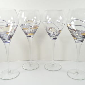 May include: Four clear wine glasses with long stems and round bases. Each glass features a decorative design of swirling blue, gold, and silver lines on the bowl. The glasses are arranged in a row against a white background.