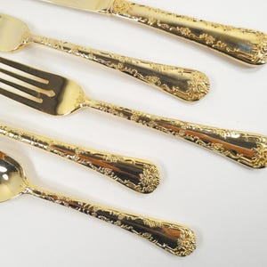 May include: Close-up of gold-colored flatware set, including a knife, forks, and a spoon. The utensils have ornate floral detailing along the handles and are arranged diagonally. The set is likely for formal dining.