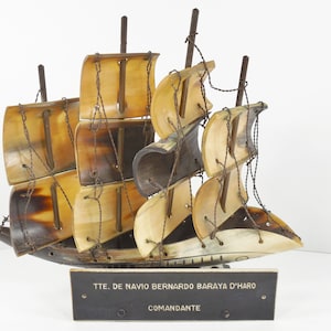 May include: A wooden ship model with sails made of carved horn. The ship is mounted on a wooden base with a plaque that reads "TTE. DE NAVIO BERNARDO BARAYA D'HARO COMANDANTE".