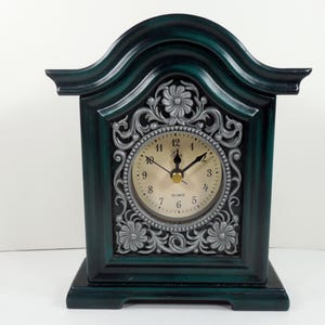 May include: A dark teal quartz clock with a decorative silver floral design around the clock face. The clock has a curved top and a rectangular base. The clock hands are black and the numbers are in a classic font.