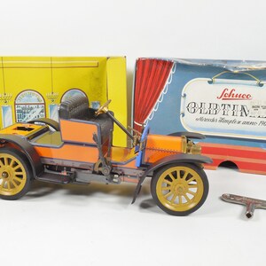 May include: A vintage orange and black toy car with a key wind-up mechanism. The car is labeled "Schuco Oldtimer Mercedes Simplex anno 1902".