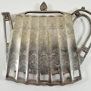 May include: Silver-colored metal teapot-shaped trivet with a decorative floral pattern. The trivet has a handle and a spout, and is designed to protect surfaces from hot teapots. The trivet measures approximately 10 inches long and 8 inches wide.