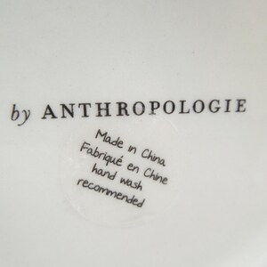 May include: Close-up of a white ceramic surface with the text "by ANTHROPOLOGIE" in black. A circular sticker reads "Made in China, Fabriqu&eacute; en Chine, hand wash recommended."
