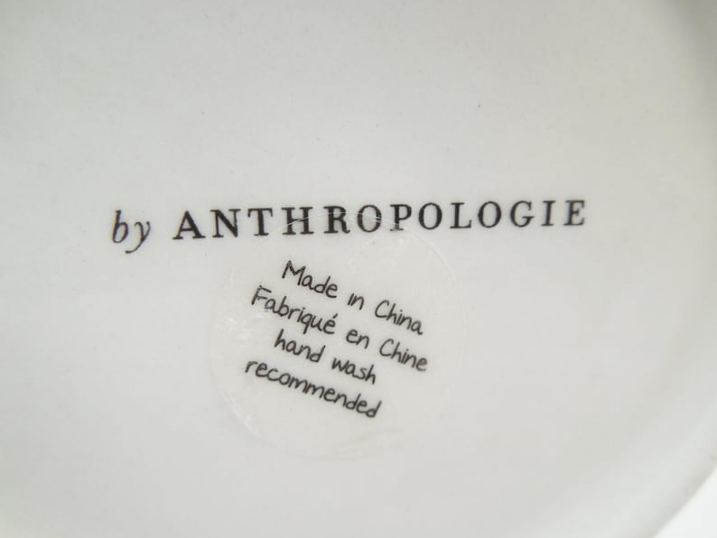 May include: Close-up of a white ceramic surface with the text "by ANTHROPOLOGIE" in black. A circular sticker reads "Made in China, Fabriqu&eacute; en Chine, hand wash recommended."