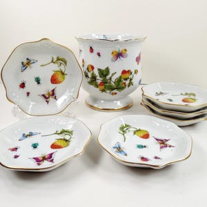 May include: A white porcelain set consisting of one pedestal vase and five small scalloped dishes. Each piece features hand-painted motifs of red strawberries, green leaves, colorful butterflies, and small insects, all finished with a gold rim.