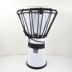 May include: A white Toca djembe drum with black rope detailing. The drum has a white drumhead and a flared base. The black rope is used to tune the drum. The drum is designed for percussion and music.