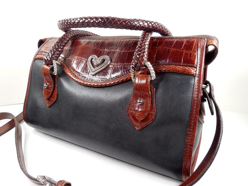 May include: A structured handbag featuring a smooth black leather body contrasted with dark brown crocodile-embossed leather trim. It includes braided leather handles, a silver-toned heart-shaped metal emblem, and a detachable shoulder strap.