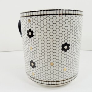 May include: White ceramic mug with a black and white honeycomb pattern, black flower accents, and gold dots. The mug has a black handle and a black border at the top and bottom. A decorative kitchen item.