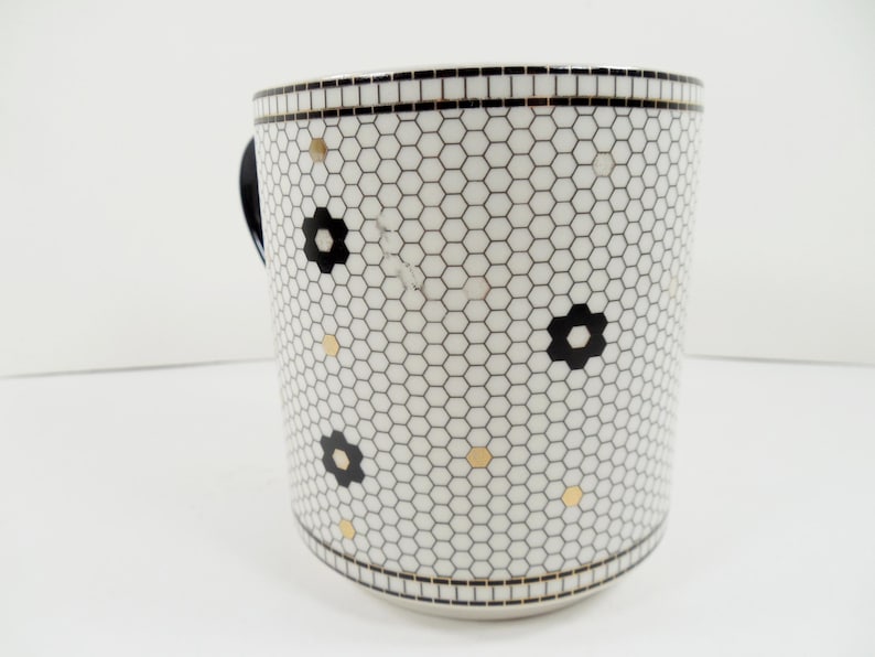 May include: White ceramic mug with a black and white honeycomb pattern, black flower accents, and gold dots. The mug has a black handle and a black border at the top and bottom. A decorative kitchen item.