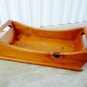 May include: A wooden tray with a natural finish. The tray has a rectangular shape with curved sides and integrated handles. The wood grain is visible, and the tray appears to be handcrafted. The tray is suitable for serving or display.