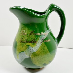 May include: Green ceramic pitcher with a curved handle and spout. The pitcher has a glossy finish with a design of yellow and white accents. Gold text on the front reads "The Hermitage Nashville Tenn."