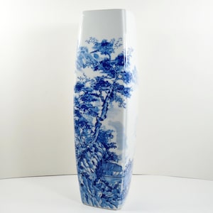 May include: Tall, rectangular white vase with a blue landscape design. The blue design depicts trees, mountains, and a small structure, creating a serene, nature-inspired aesthetic. The vase has a unique, elongated shape.