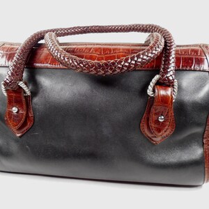 May include: A structured handbag featuring a smooth black leather body contrasted with a dark brown crocodile-embossed leather top panel. It includes two braided dark brown leather handles attached with silver-toned metal hardware and rivets.