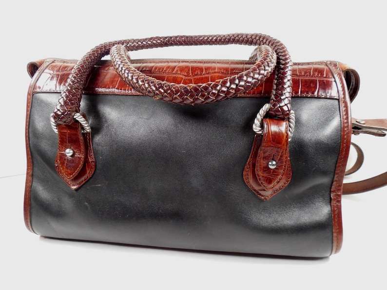 May include: A structured handbag featuring a smooth black leather body contrasted with a dark brown crocodile-embossed leather top panel. It includes two braided dark brown leather handles attached with silver-toned metal hardware and rivets.