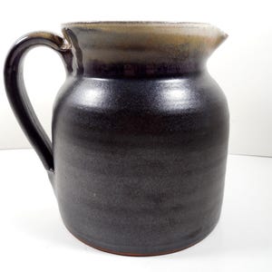 May include: A handmade ceramic pitcher with a dark, matte finish. The pitcher has a large handle and a spout for easy pouring. The rim has a lighter, contrasting color. A functional and decorative piece for the kitchen or dining room.