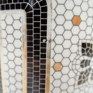 May include: Close-up of a white ceramic object with a black and gold mosaic design. The design features a honeycomb pattern, black squares, and gold accents. A decorative floral motif is also visible.