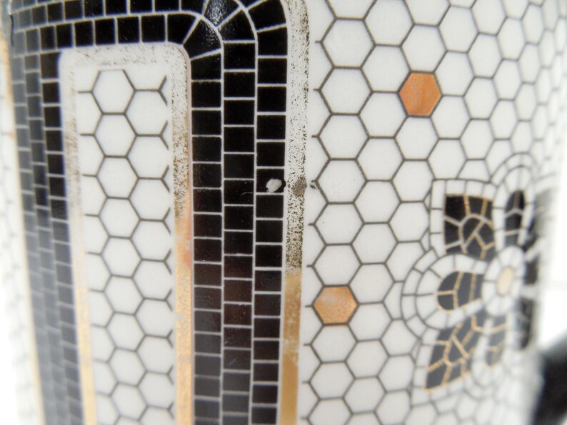 May include: Close-up of a white ceramic object with a black and gold mosaic design. The design features a honeycomb pattern, black squares, and gold accents. A decorative floral motif is also visible.