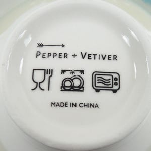 May include: A close-up of the base of a white ceramic bowl. The bowl's base features the text "PEPPER + VETIVER" and black symbols indicating dishwasher, freezer, and microwave safe. The text "MADE IN CHINA" is also present.