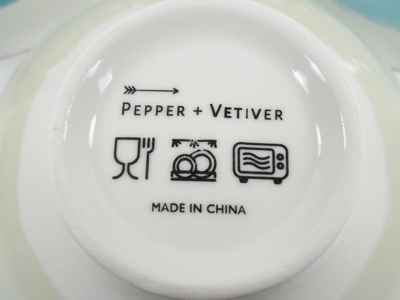 May include: A close-up of the base of a white ceramic bowl. The bowl's base features the text "PEPPER + VETIVER" and black symbols indicating dishwasher, freezer, and microwave safe. The text "MADE IN CHINA" is also present.