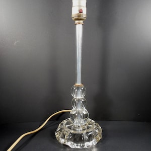 May include: A clear glass table lamp base featuring a tall, slender stem rising from a stack of three spherical glass accents. The base is circular with a scalloped edge, and the top includes a metal socket with a small red switch component.