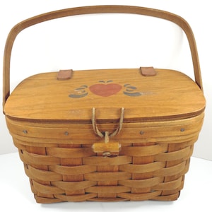 May include: A light brown woven basket with a wooden handle and lid. The lid features a painted red heart with blue accents. The basket has a wooden latch and is suitable for storage or display.