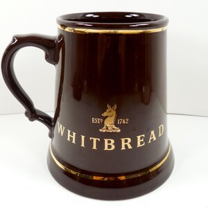 May include: A dark brown Whitbread beer mug with a gold rim and handle. The mug features the Whitbread logo and the text "WHITBREAD" in gold lettering. The mug has a capacity of approximately 16 ounces.