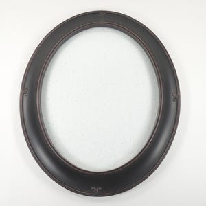 May include: Oval-shaped, dark brown picture frame with decorative flourishes at the top, bottom, and sides. The frame has a smooth, rounded profile and a lighter brown inner edge. The center is empty, ready for a photo or artwork.