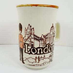 May include: Cream-colored ceramic mug featuring a brown illustration of London landmarks, including Tower Bridge and a Beefeater guard. The word "London" is printed in a bold, brown font. The rim of the mug has a brown trim.